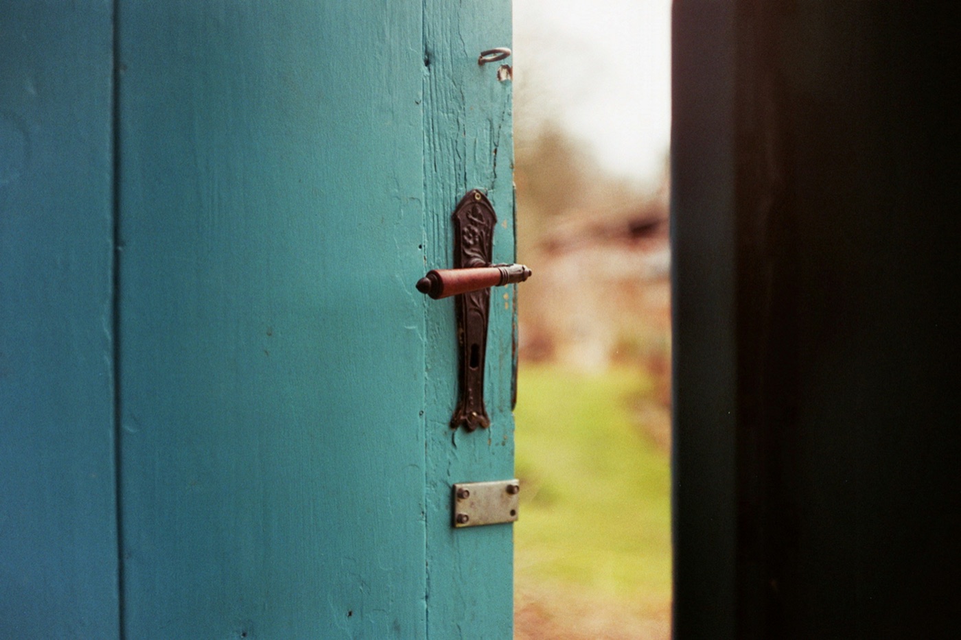 A teal wooden door standing ajar, a hand's reach from the handle, with a sunlit garden beyond